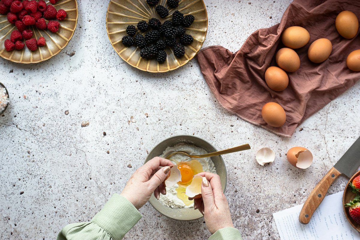 Mains cassant un œuf dans un bol de farine, œufs et petits fruits autour